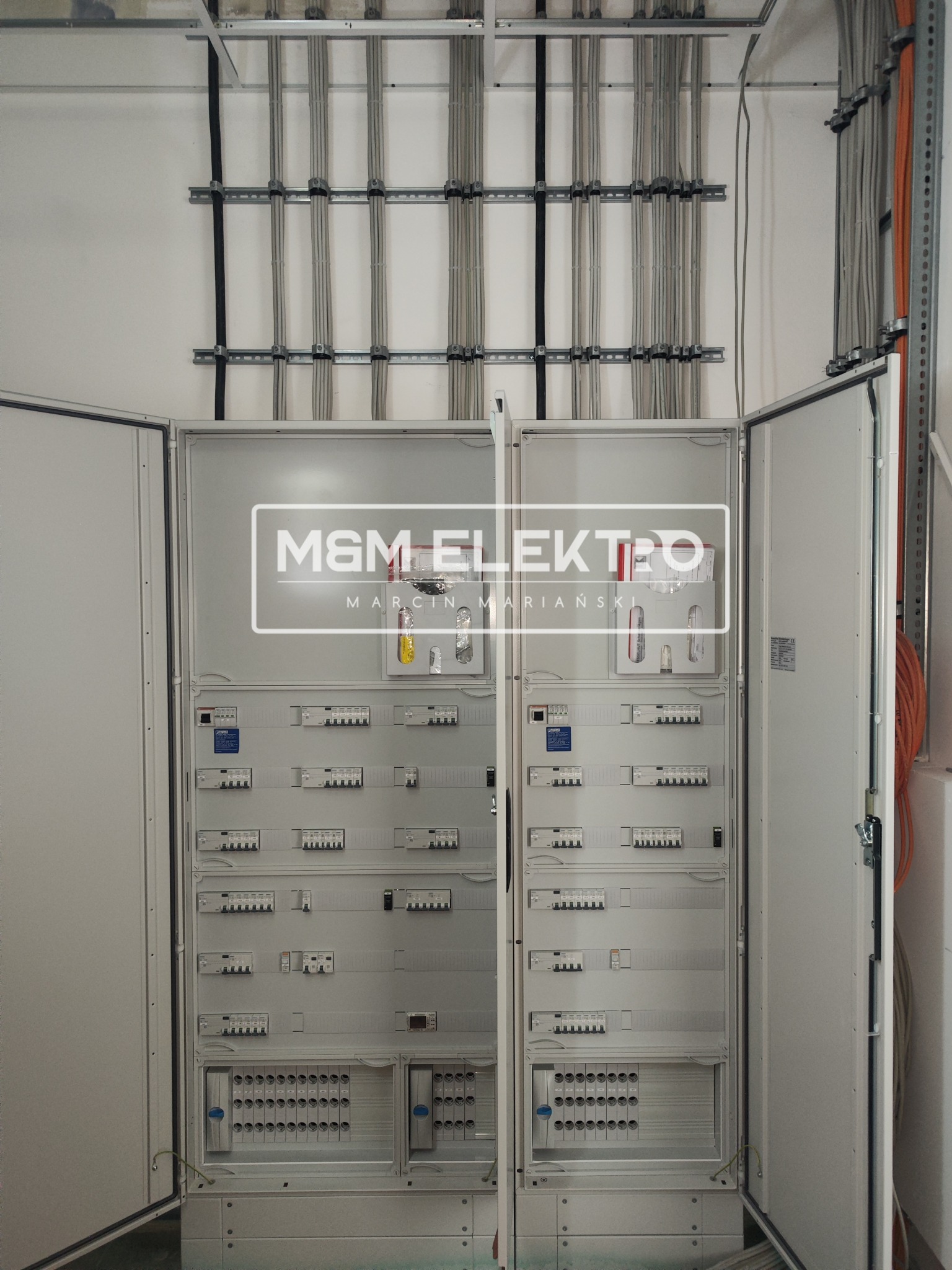 Otwarta szafa elektryczna z zamontowanymi zabezpieczeniami i przewodami, widoczne szyny montażowe na ścianie i logo firmy M&M Elektro.