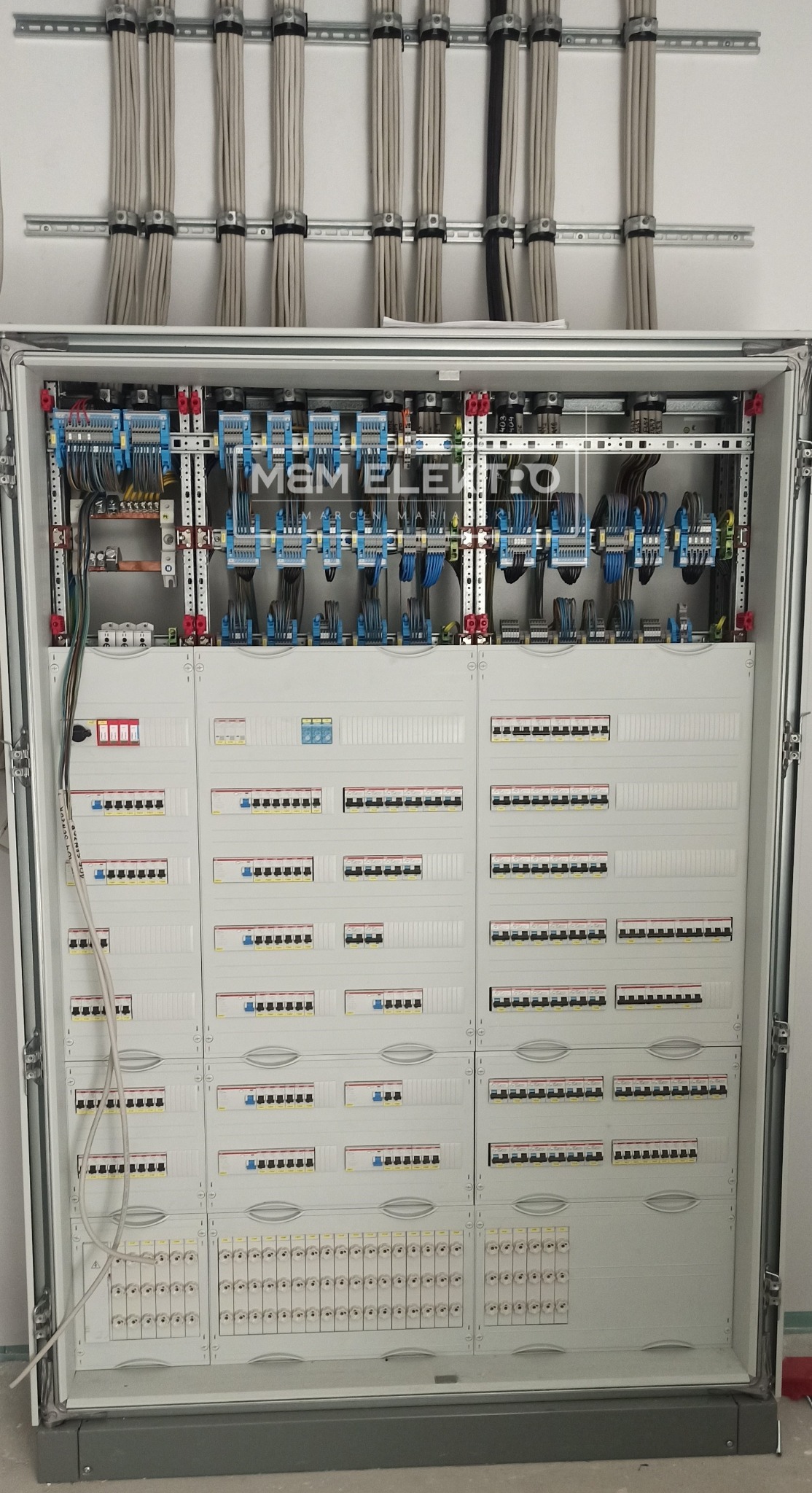 Otwarta szafa rozdzielcza z licznymi bezpiecznikami, okablowaniem i listwami montażowymi, widoczne uporządkowane kable w górnej części szafy.