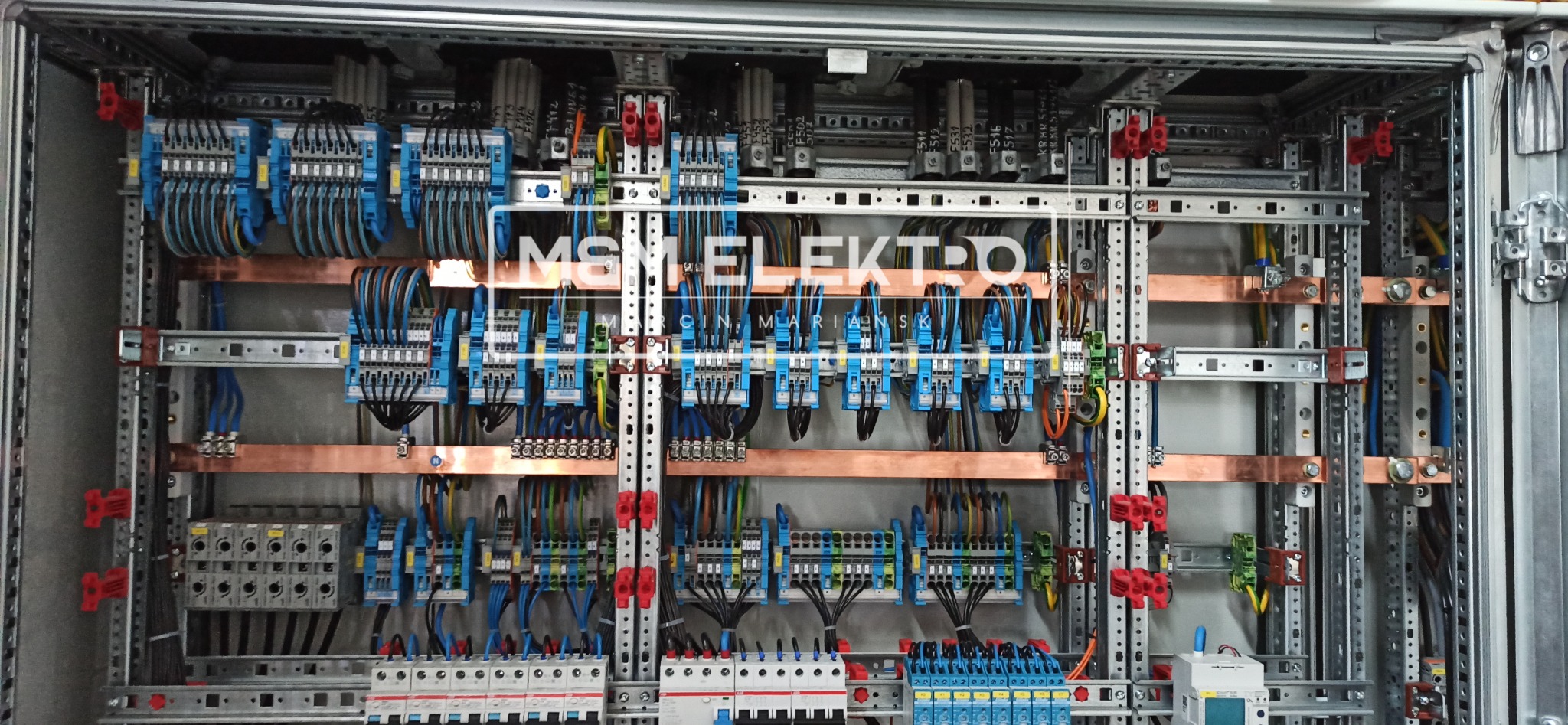 Rozbudowana skrzynka elektryczna z licznymi niebieskimi złączkami, miedzianymi szynami i przewodami w różnych kolorach, zamontowana w metalowej obudowie z widocznymi elementami mocującymi.