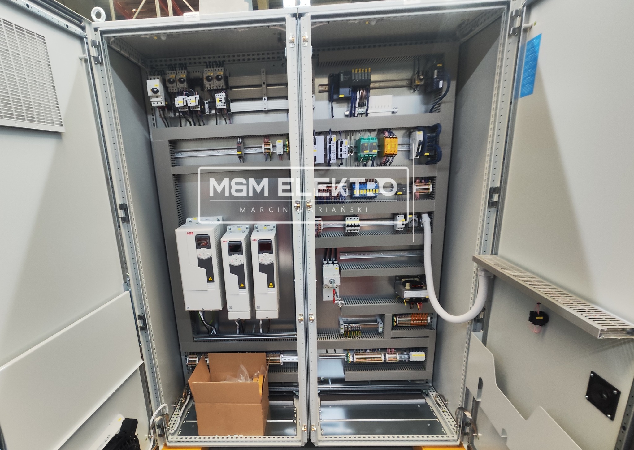 Otwarta szafa sterownicza z trzema falownikami ABB po lewej stronie i różnorodną aparaturą elektryczną po prawej, widoczna kartonowa skrzynka na dole po lewej.