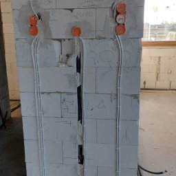 J&L - Instalacja elektryczna w surowym stanie: białe przewody prowadzone po ścianie z bloczków, pomarańczowe puszki instalacyjne, widoczne napisy na ścianie, peszle na podłodze.