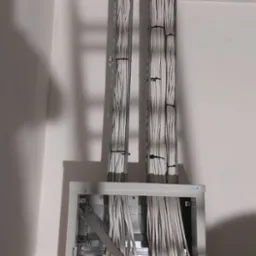 Pionowe wiązki kabli elektrycznych, zabezpieczone czarnymi opaskami, prowadzone w metalowych korytkach od sufitu podwieszanego do otwartej szafy rozdzielczej na ścianie.