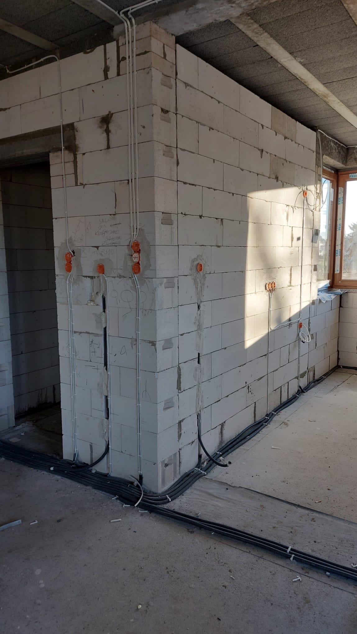 Surowy stan budynku z widocznymi przewodami elektrycznymi w peszlach przy podłodze i puszkami instalacyjnymi w ścianach z betonu komórkowego.