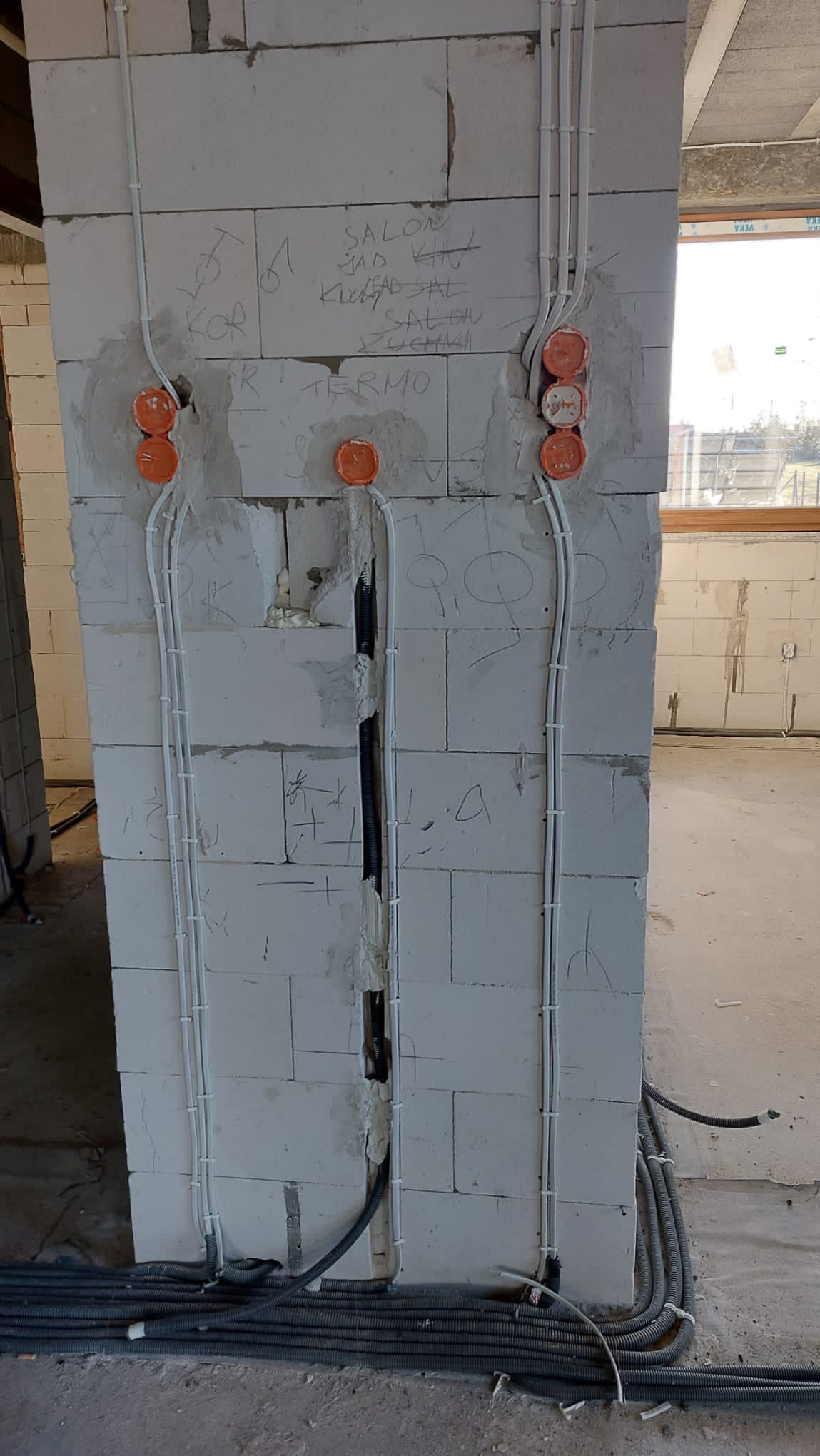 Instalacja elektryczna w surowym stanie: białe przewody prowadzone po ścianie z bloczków, pomarańczowe puszki instalacyjne, widoczne napisy na ścianie, peszle na podłodze.