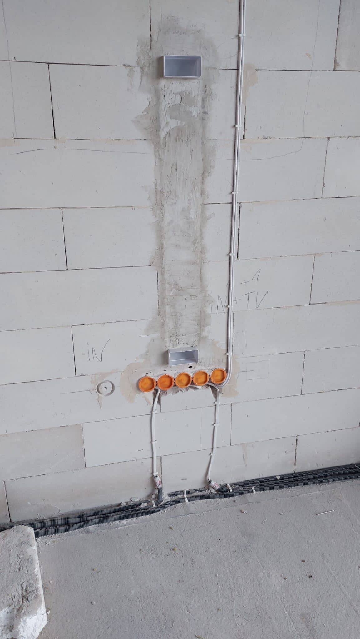 Surowa ściana z betonu komórkowego z wyfrezowanymi bruzdami i zamontowanymi puszkami elektrycznymi w kolorze pomarańczowym, widoczne kable w peszlach i rurkach PCV, przygotowane do dalszych prac...