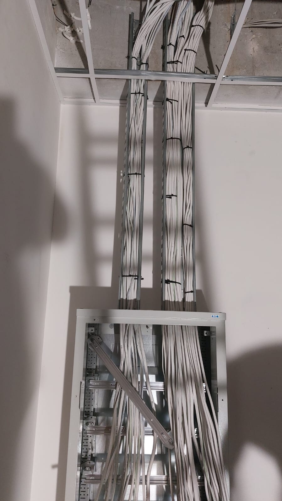 Pionowe wiązki kabli elektrycznych, zabezpieczone czarnymi opaskami, prowadzone w metalowych korytkach od sufitu podwieszanego do otwartej szafy rozdzielczej na ścianie.