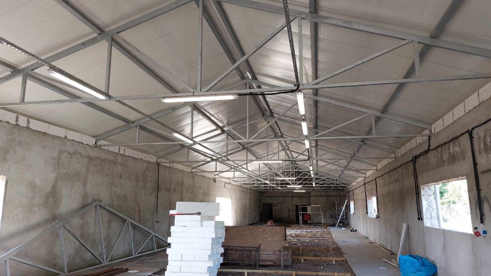 Wnętrze hali w trakcie budowy z widoczną konstrukcją dachu, oświetleniem liniowym LED i instalacją elektryczną na ścianach.