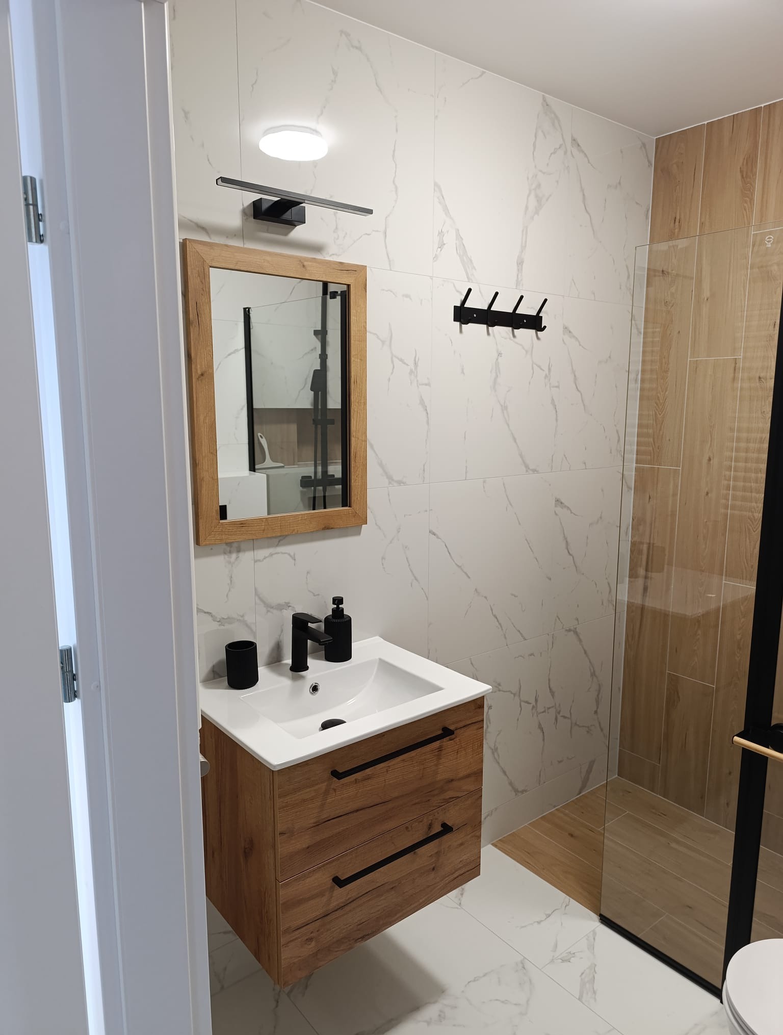 Nowoczesna łazienka po remoncie: białe marmurowe płytki, drewniane akcenty, czarna armatura i kabina prysznicowa z drewnianymi panelami. Minimalistyczny design.