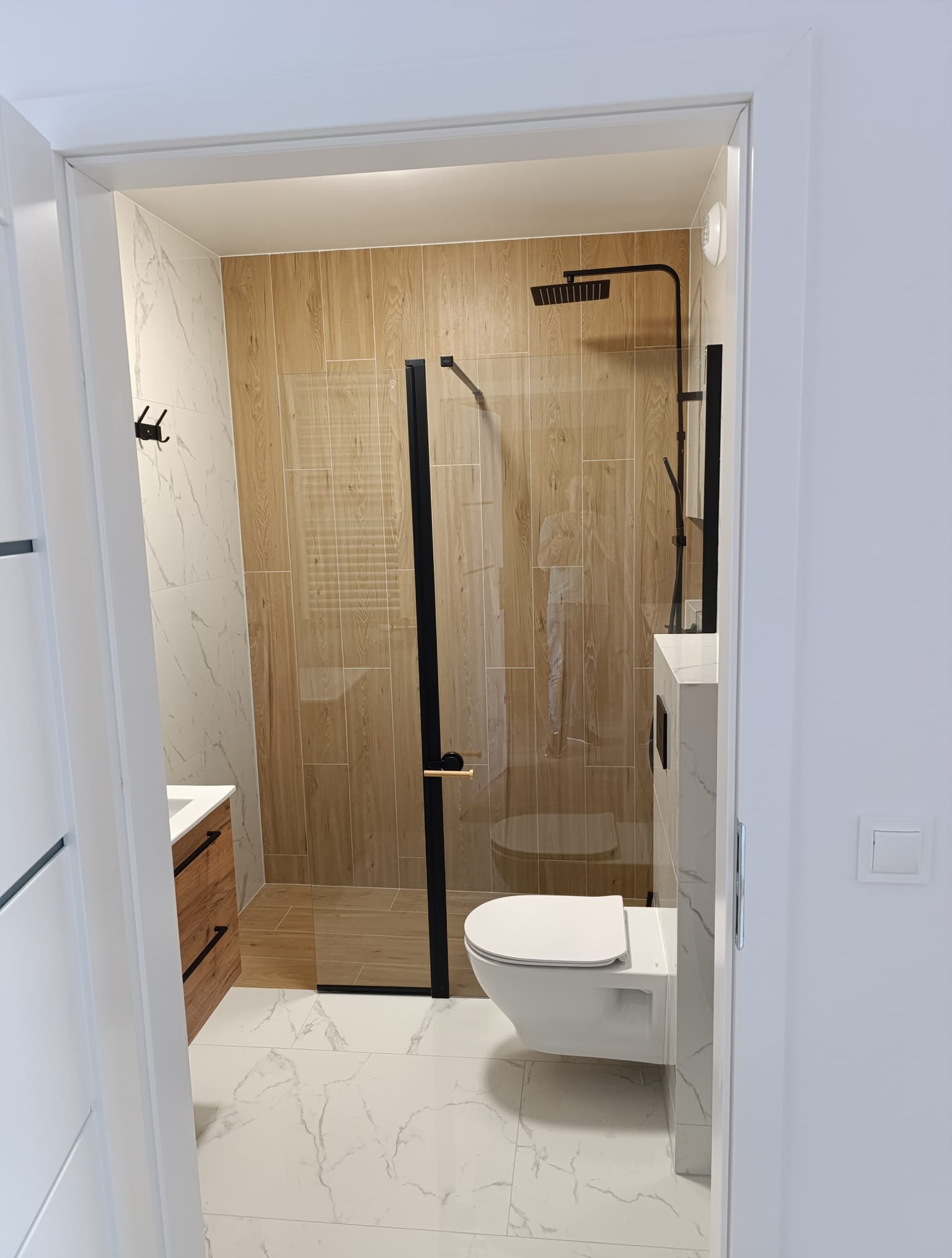 Nowoczesna łazienka z kabiną prysznicową typu walk-in, wykończona płytkami imitującymi drewno i marmur. Czarna armatura i minimalistyczne dodatki.