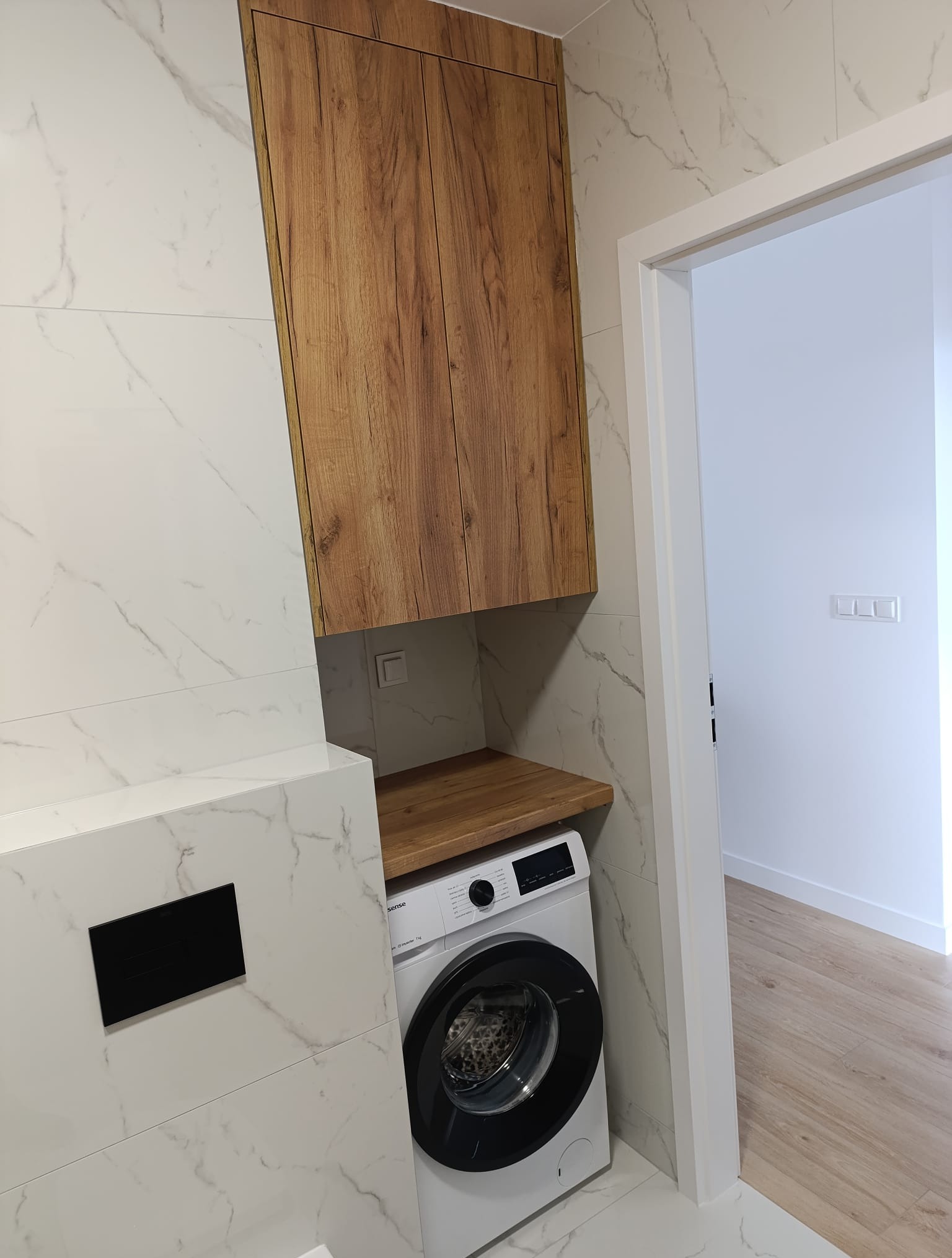 Nowoczesna łazienka z marmurowymi płytkami, drewnianą szafką nad pralką i blatem z drewna. Widoczny fragment podłogi z paneli i białe drzwi.
