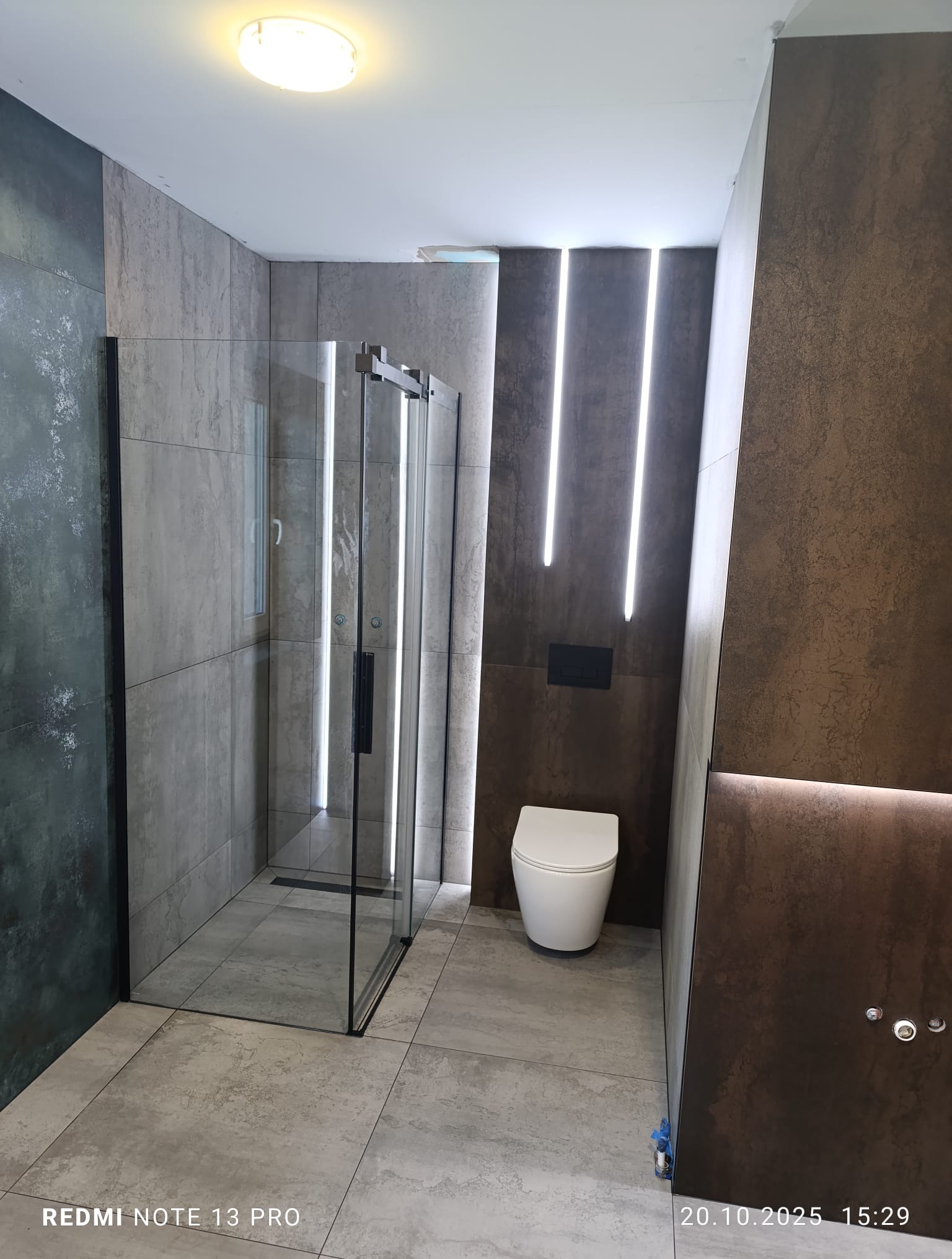 Nowoczesna łazienka z kabiną prysznicową typu walk-in, wykończona płytkami w odcieniach szarości i brązu. Uwagę zwracają pionowe pasy świetlne na ścianie.