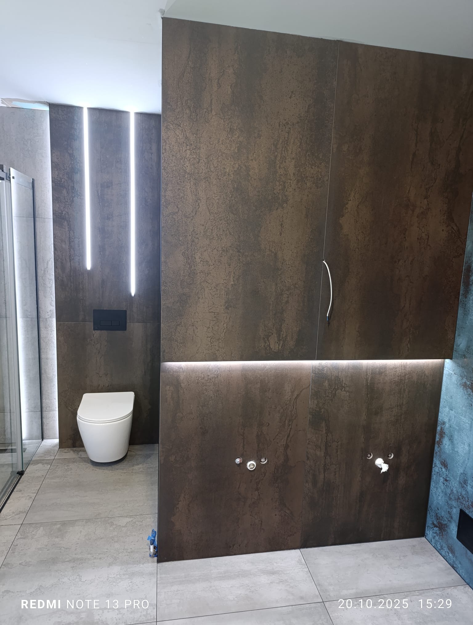 Nowoczesna łazienka z brązowymi płytkami imitującymi kamień, podświetleniem LED i wiszącą toaletą. Widoczne przyłącza wodne i szklana kabina prysznicowa.