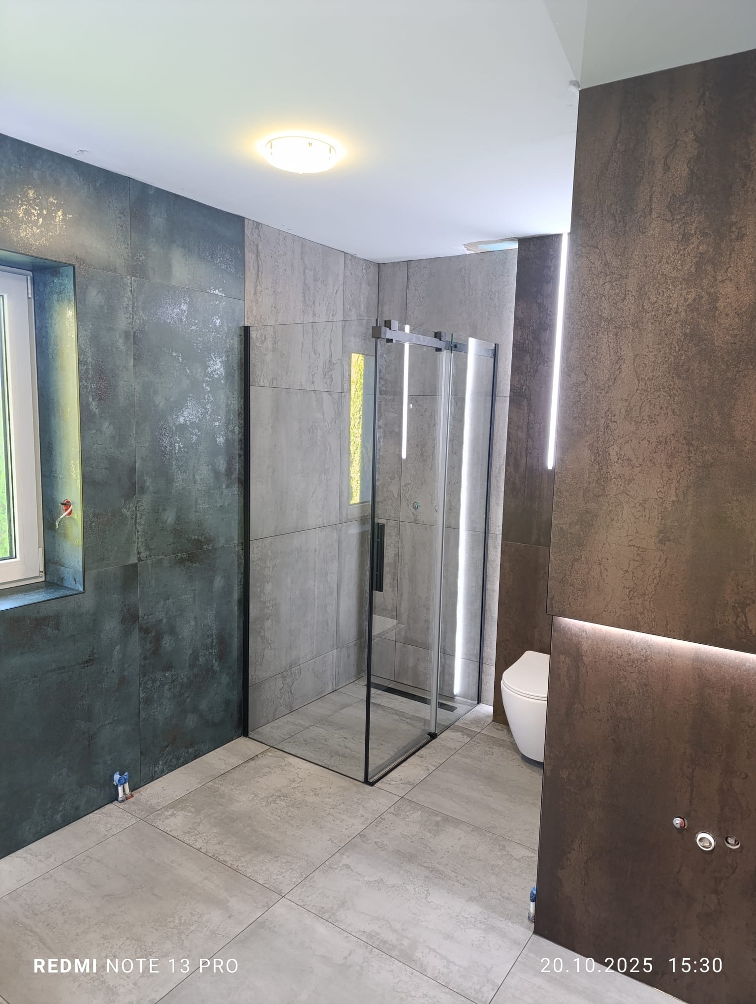 Nowoczesna łazienka z kabiną prysznicową typu walk-in, wykończona płytkami w odcieniach szarości i brązu, z subtelnym oświetleniem LED. Widoczny fragment toalety.