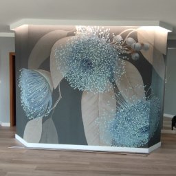 OK-BUD - Ściana z fototapetą przedstawiającą duże, błękitne kwiaty na tle brązowych liści, podświetlona od góry, z widocznymi panelami podłogowymi.