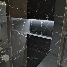 OK-BUD - Czarna marmurowa ściana z wnęką podświetloną ledami, kontrastująca z jasnymi marmurowymi płytkami podłogowymi.