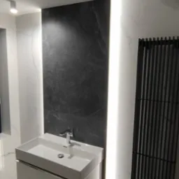 Nowoczesna łazienka z umywalką nablatową na szafce, czarnym panelem dekoracyjnym z podświetleniem LED i grzejnikiem drabinkowym w kolorze czarnym na tle białych marmurowych płytek.