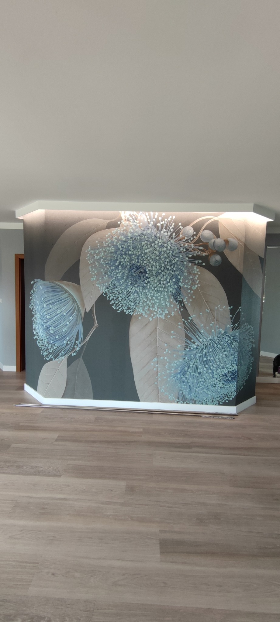 Ściana z fototapetą przedstawiającą duże, błękitne kwiaty na tle brązowych liści, podświetlona od góry, z widocznymi panelami podłogowymi.