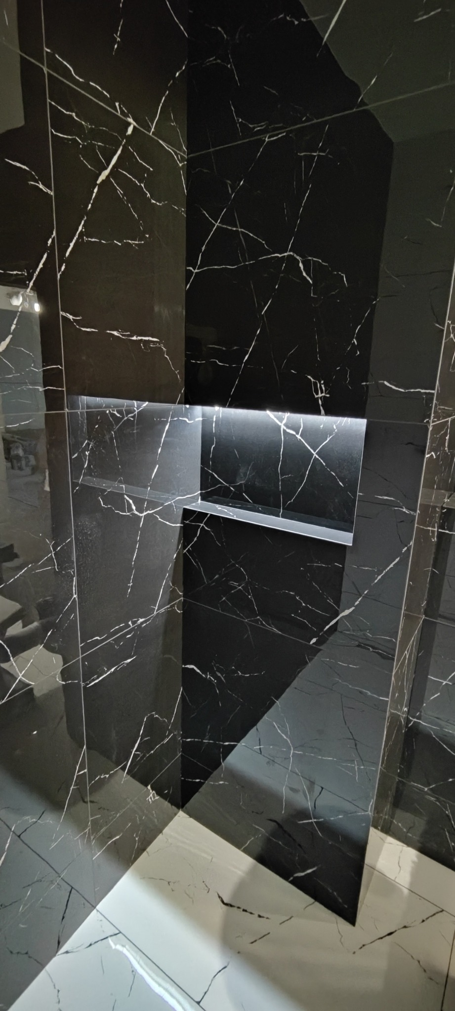 Czarna marmurowa ściana z wnęką podświetloną ledami, kontrastująca z jasnymi marmurowymi płytkami podłogowymi.