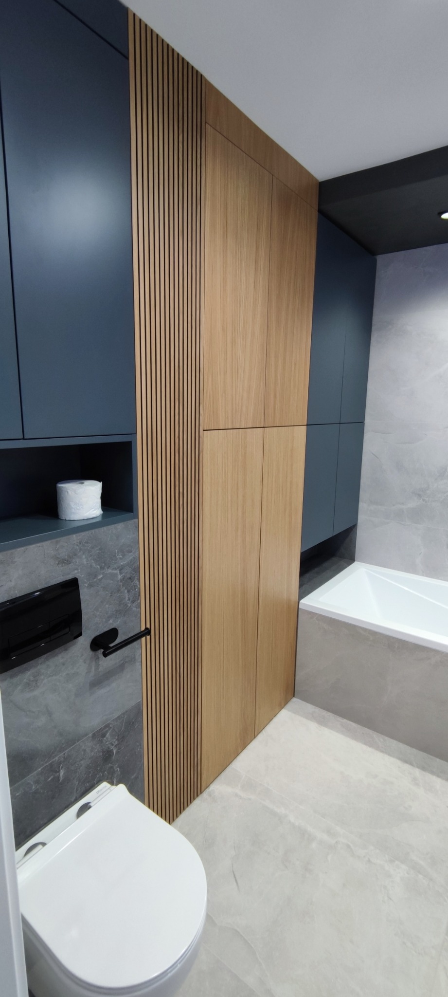 Nowoczesna łazienka z zabudową meblową w odcieniach szarości, granatu i drewna, widoczna toaleta, wanna i grzejnik drabinkowy, z rolką papieru toaletowego na półce.