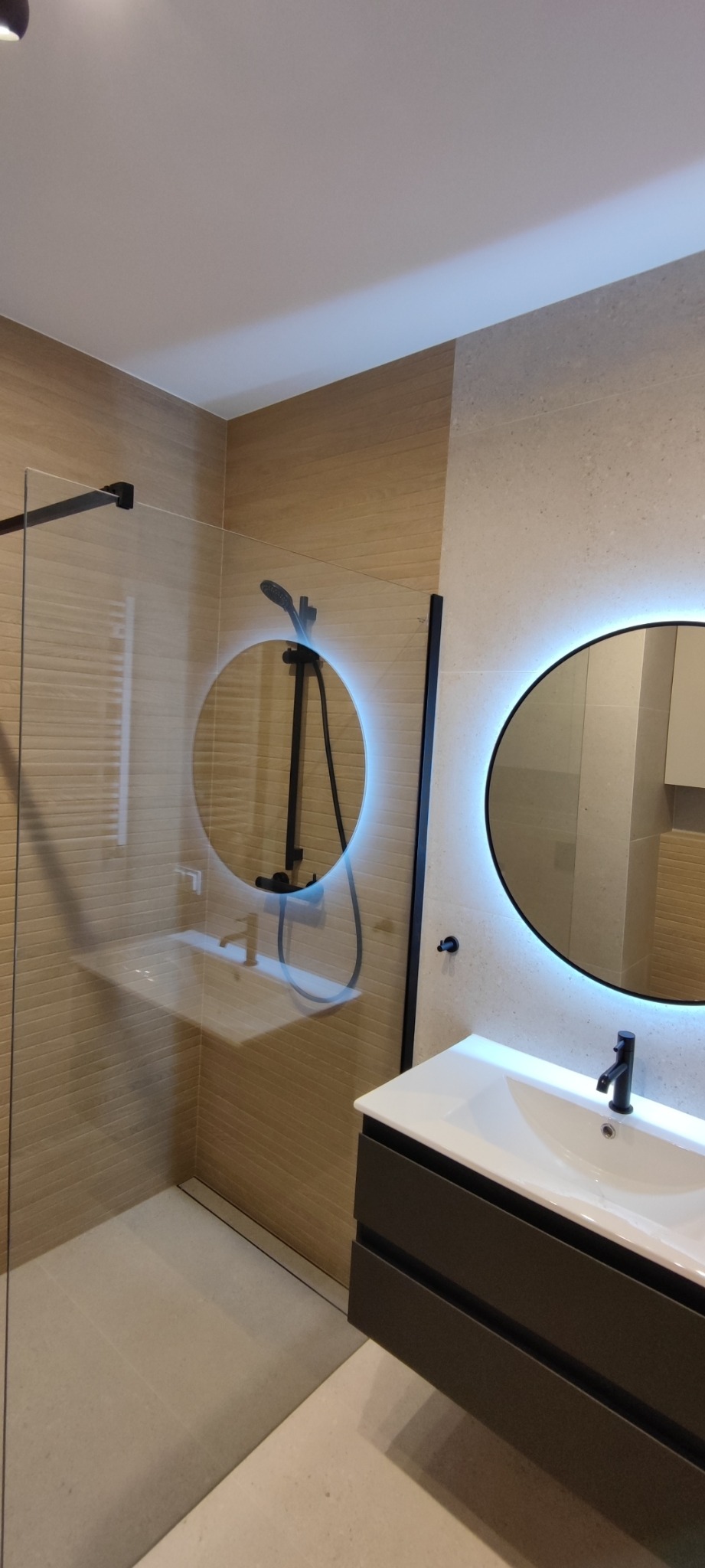 Nowoczesna łazienka z kabiną prysznicową typu walk-in, okrągłym lustrem z podświetleniem LED, umywalką nablatową na szafce w kolorze antracytowym i czarną armaturą.