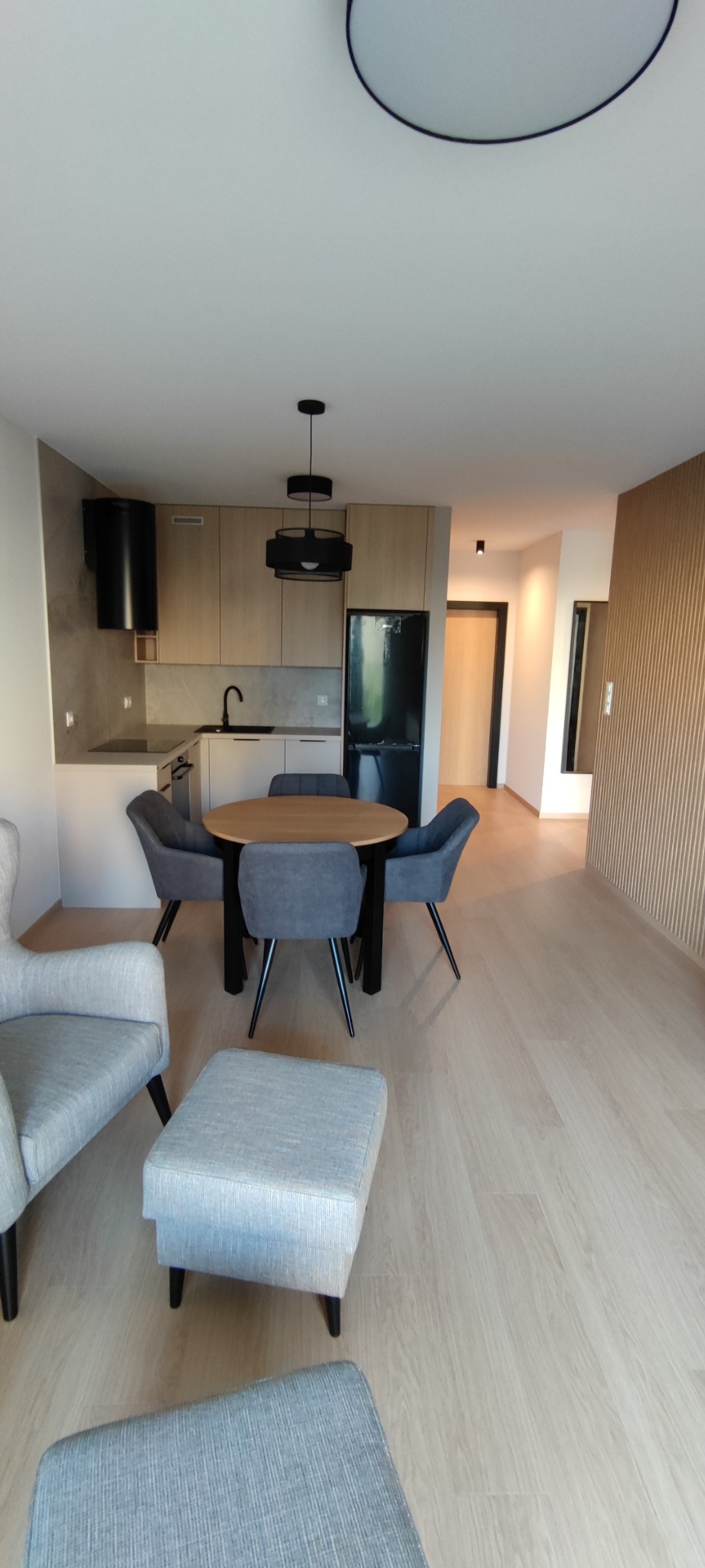 Jasne wnętrze mieszkania z aneksem kuchennym, okrągłym stołem z szarymi krzesłami, szarym fotelem z podnóżkiem i panelami na ścianie.