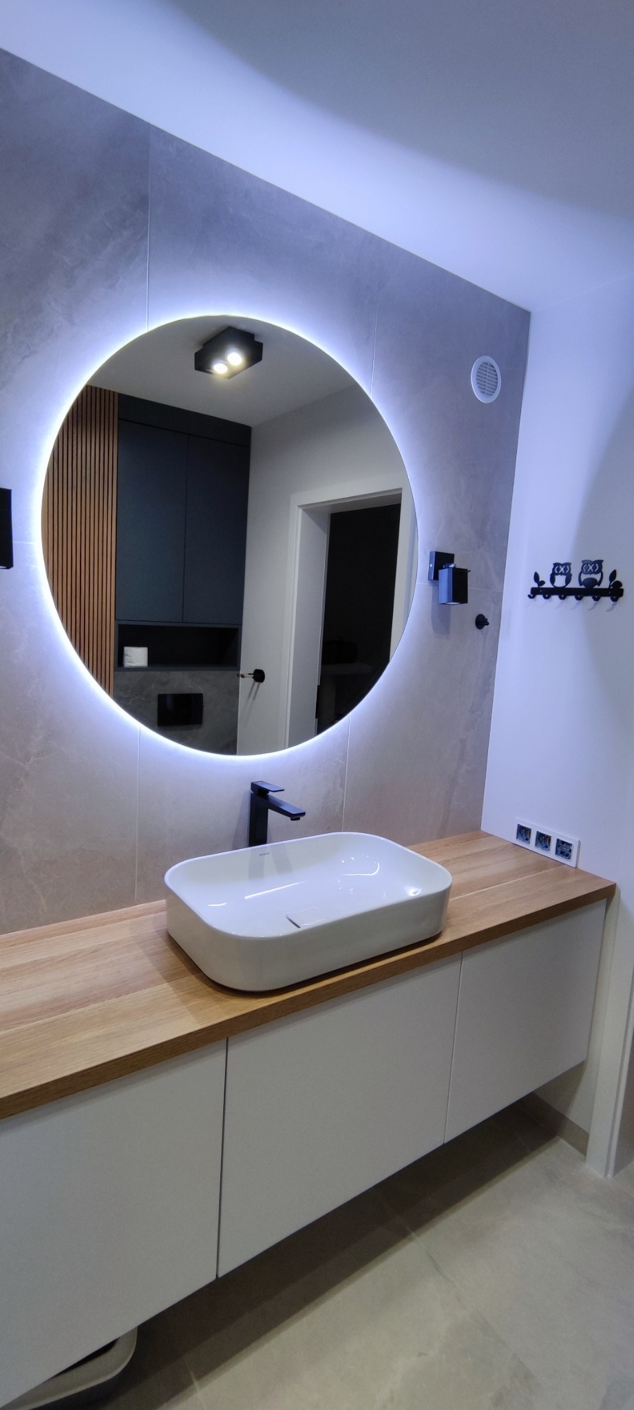 Nowoczesna łazienka z okrągłym lustrem podświetlanym LED, białą umywalką nablatową na drewnianym blacie i szarymi płytkami.