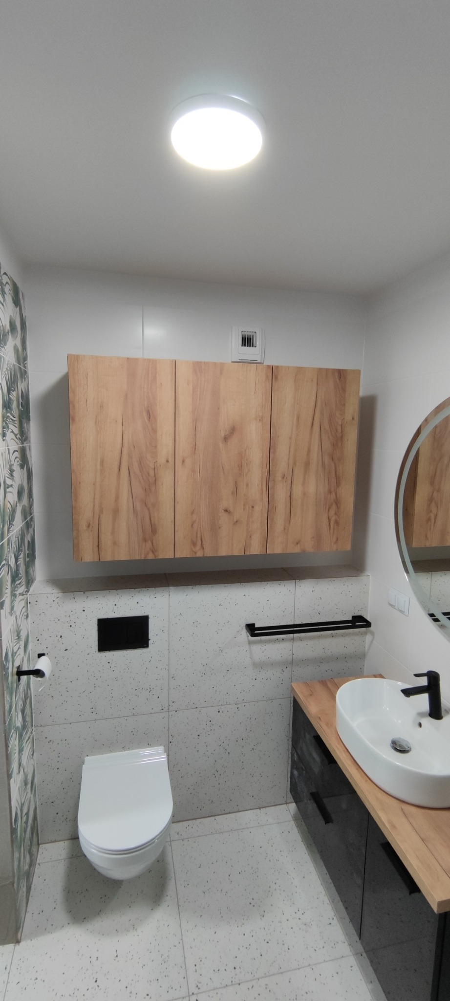 Nowoczesna łazienka z wiszącą szafką z drewnopodobnym frontem, białą umywalką nablatową z czarną armaturą, toaletą podwieszaną i płytkami w stylu lastryko.