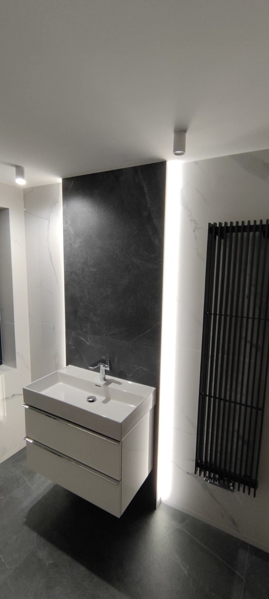 Nowoczesna łazienka z umywalką nablatową na szafce, czarnym panelem dekoracyjnym z podświetleniem LED i grzejnikiem drabinkowym w kolorze czarnym na tle białych marmurowych płytek.