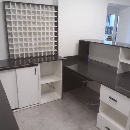 Usługowy zakład stolarski - Nowoczesna lada recepcyjna z czarnym blatem i białymi szafkami, z modułową półką na ścianie, w surowym, minimalistycznym wnętrzu.