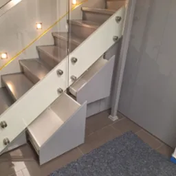 Nowoczesne schody z szarymi stopniami, szklaną balustradą mocowaną na metalowych elementach i wbudowanymi szufladami w kolorze szarym, dwie z nich są wysunięte.