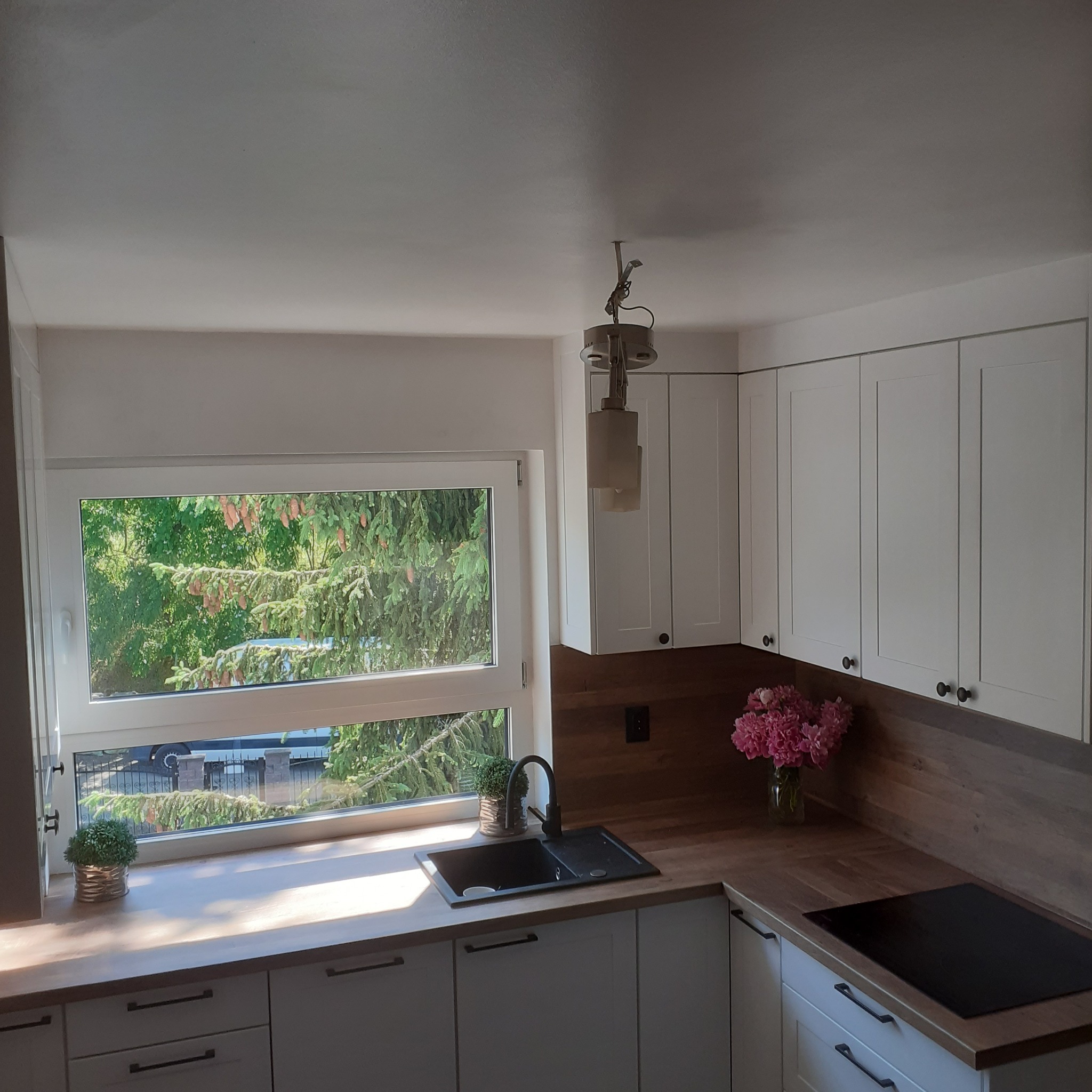 Jasna kuchnia z białymi szafkami, drewnianym blatem i widokiem na zieleń za oknem. Na blacie stoi wazon z różowymi kwiatami i czarna płyta indukcyjna.