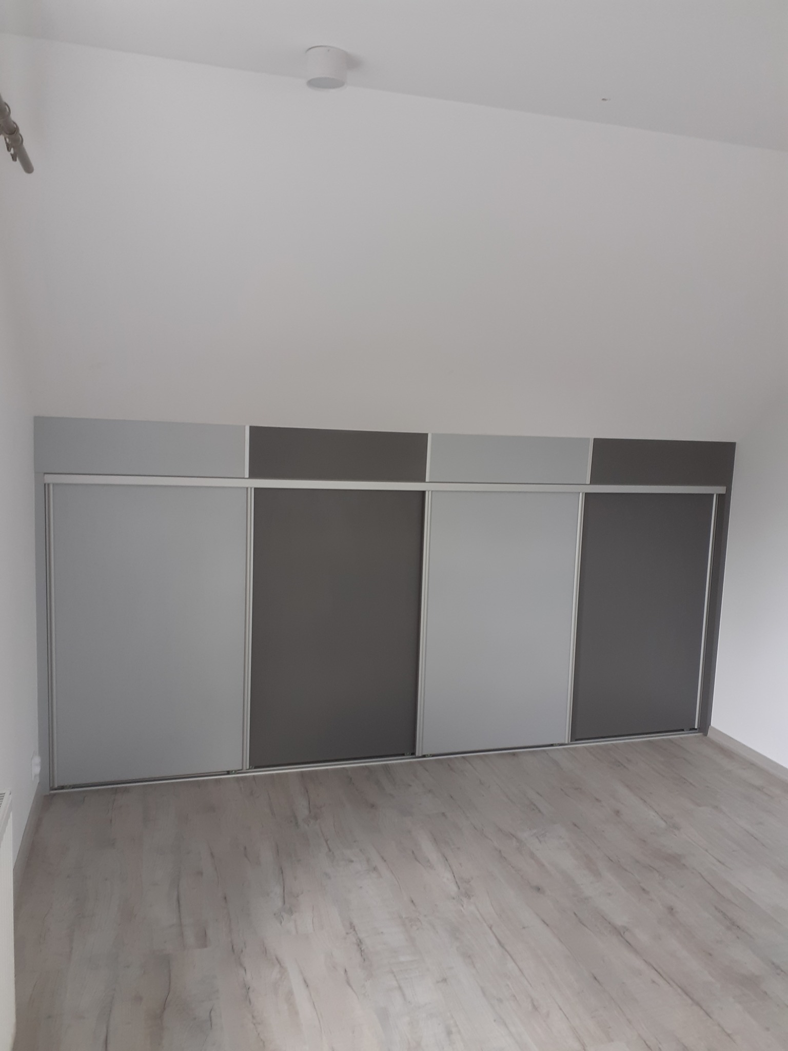 Czterodrzwiowa szafa wnękowa z przesuwnymi drzwiami w odcieniach szarości, zamontowana w pokoju na poddaszu ze skośnym sufitem i drewnianą podłogą.