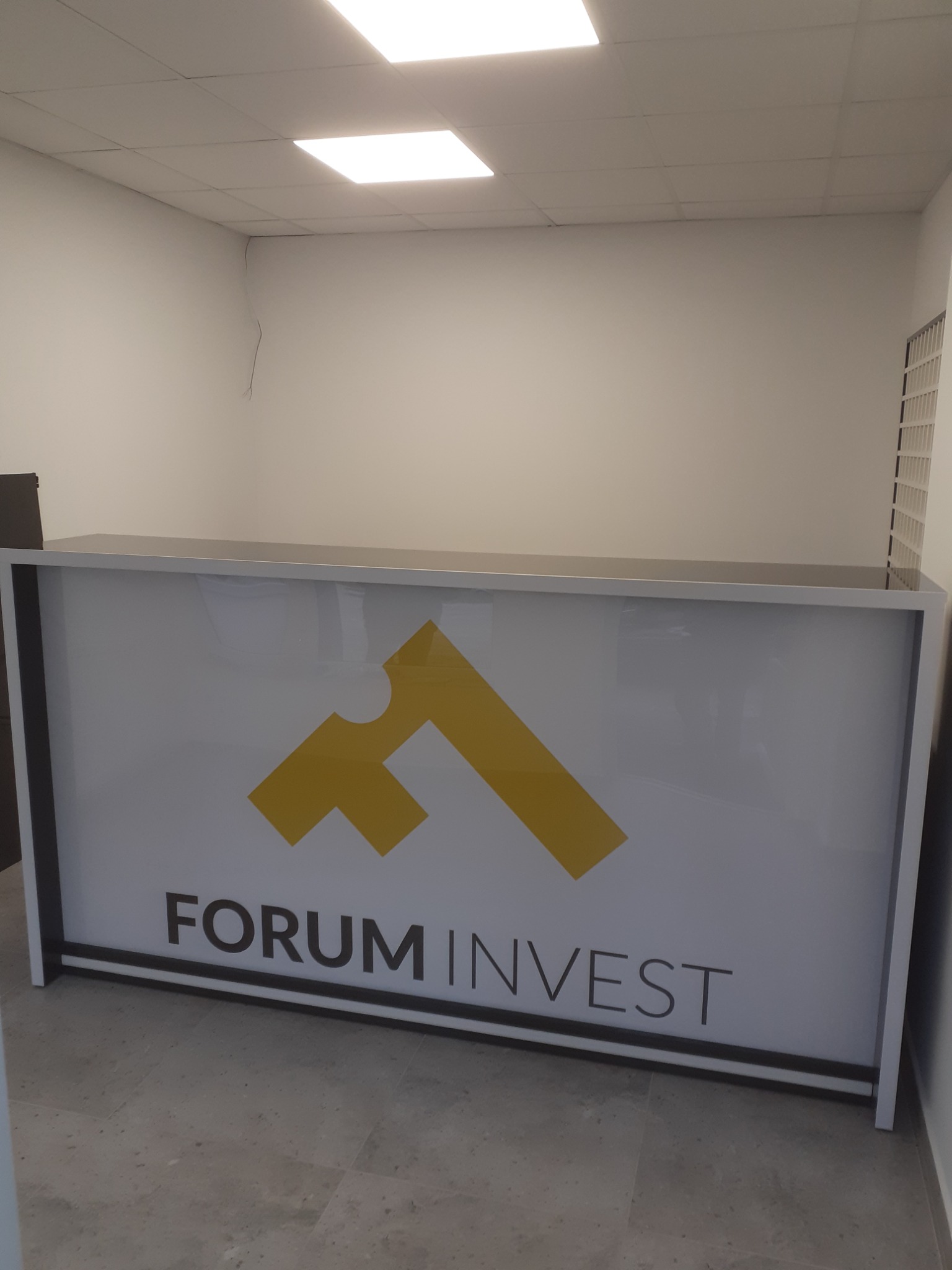 Biała lada recepcyjna z logo Forum Invest w minimalistycznym wnętrzu biurowym, oświetlona lampą sufitową.