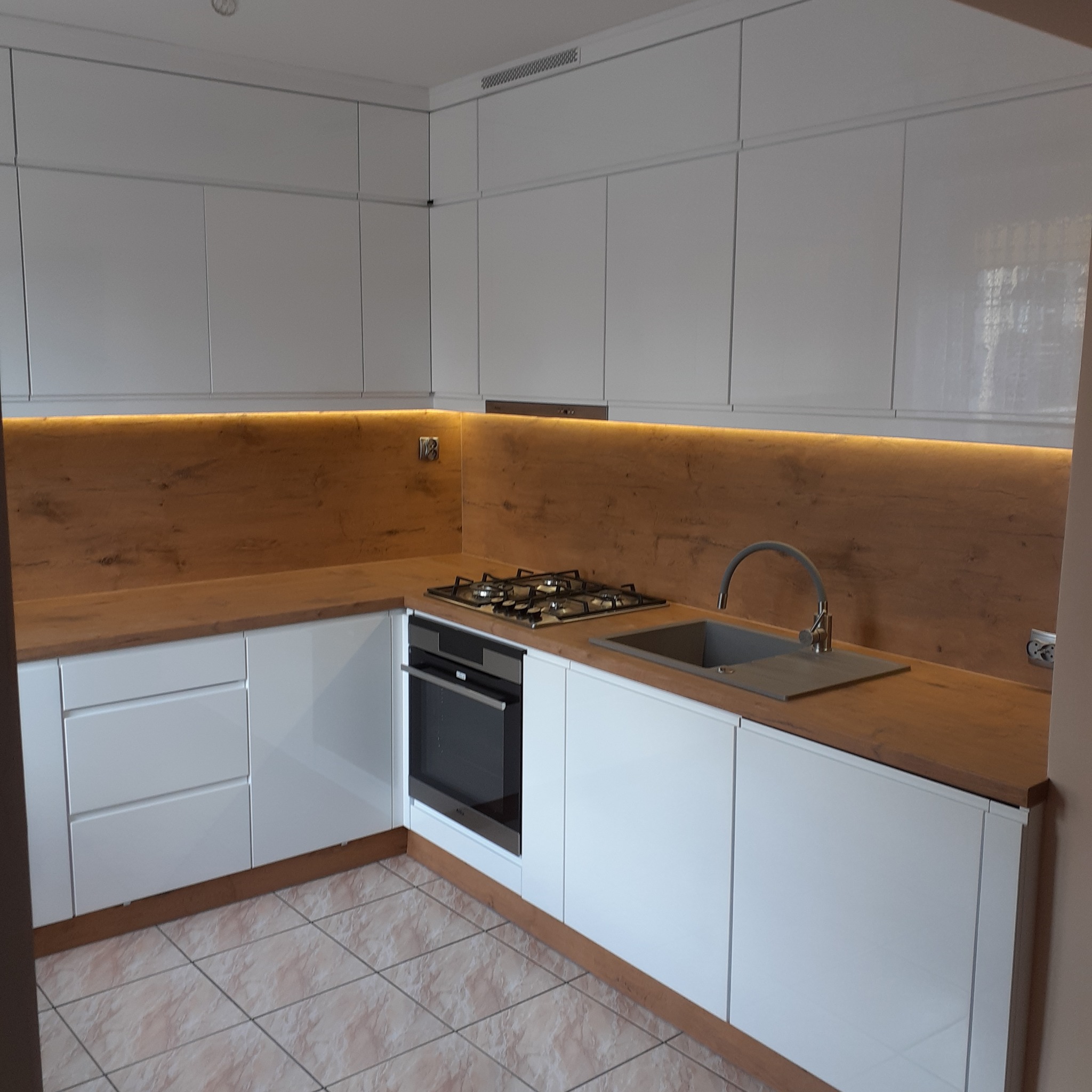 Nowoczesna kuchnia z białymi, lakierowanymi frontami, drewnianym blatem i podświetleniem LED nad blatem. Widoczny zlew, płyta grzewcza i piekarnik.