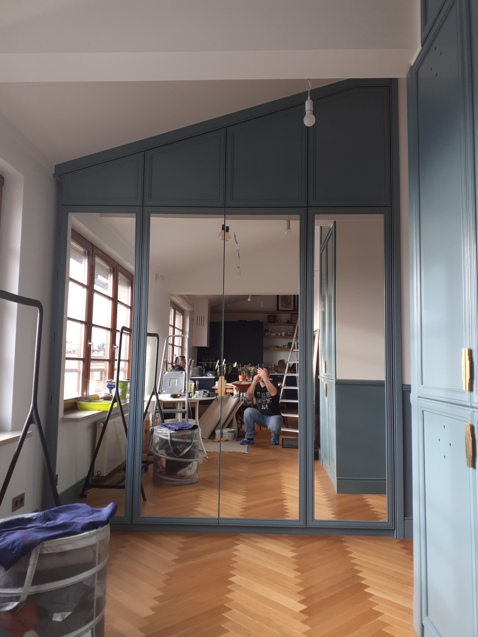 Szafa wnękowa z lustrzanymi drzwiami w kolorze szaro-niebieskim, widoczne odbicie wnętrza mieszkania podczas montażu, parkiet w jodełkę na podłodze.