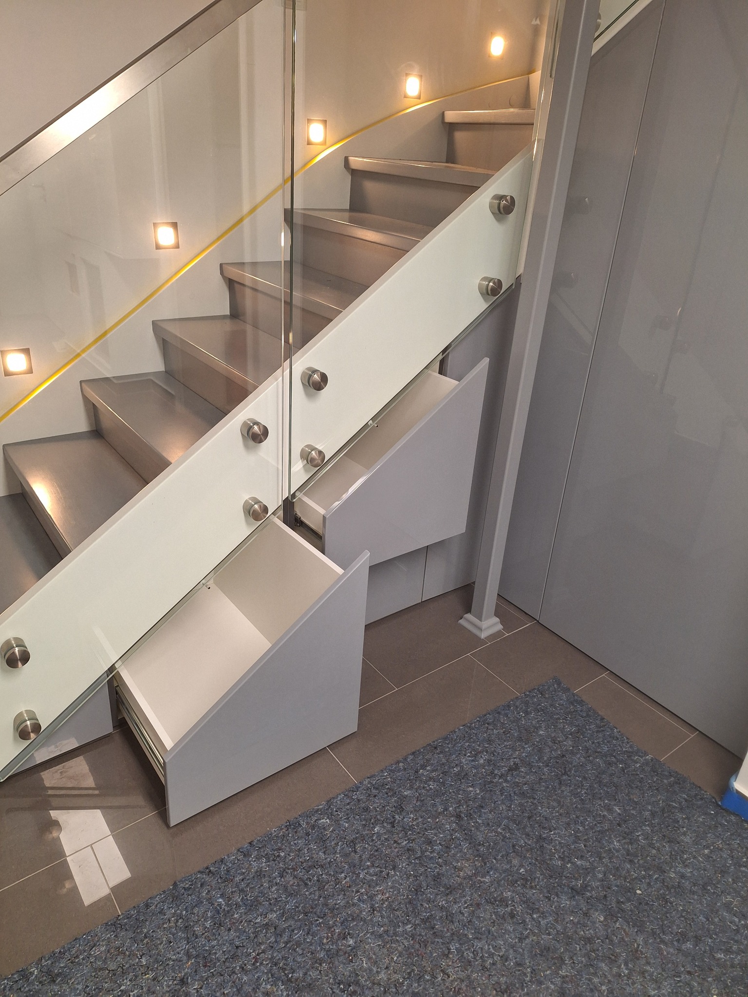 Nowoczesne schody z szarymi stopniami, szklaną balustradą mocowaną na metalowych elementach i wbudowanymi szufladami w kolorze szarym, dwie z nich są wysunięte.