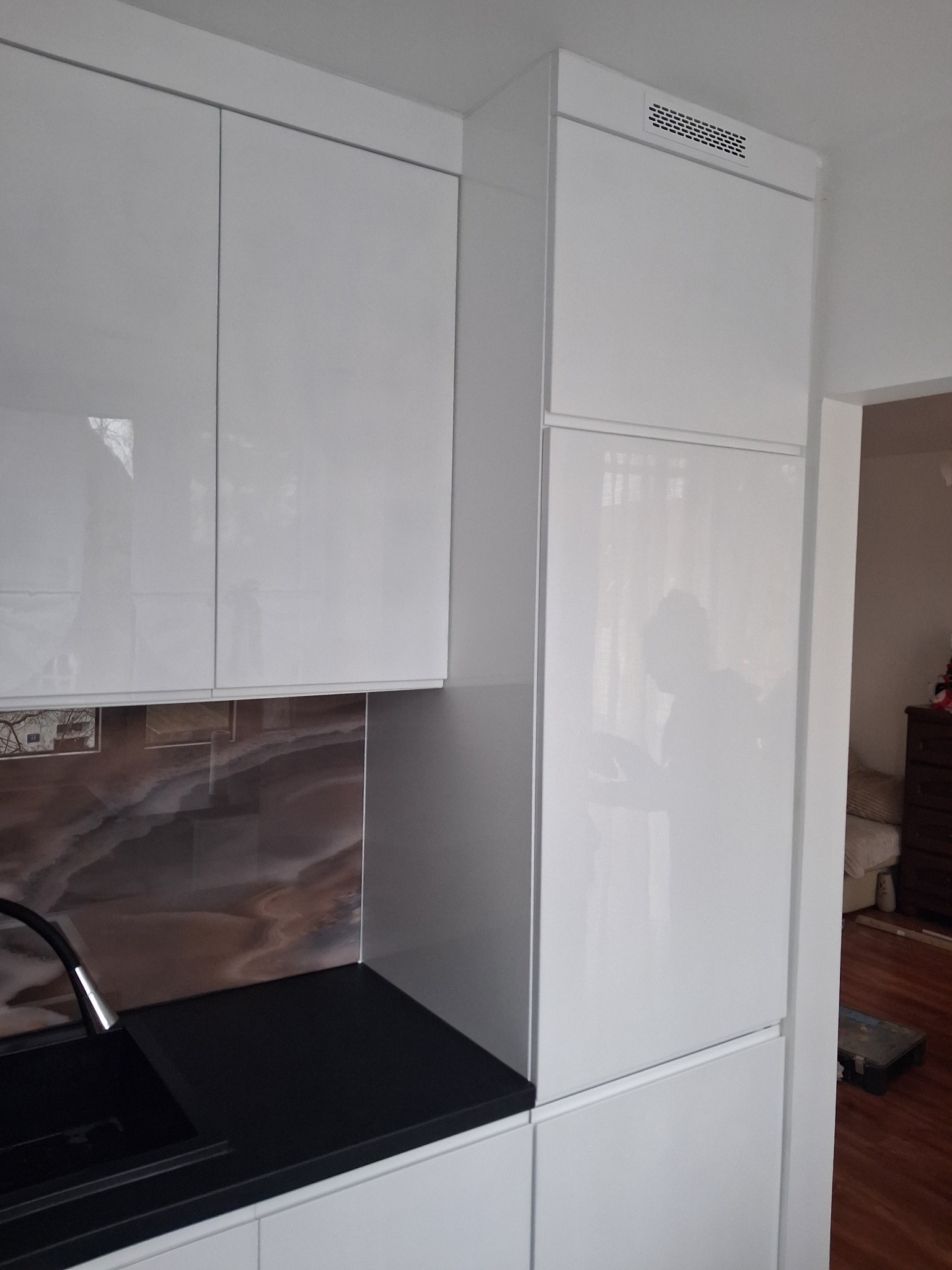 Biała, lakierowana zabudowa kuchenna z czarnym blatem i dekoracyjnym panelem ściennym. Widoczny fragment zlewu i armatury. Minimalistyczny design z elementami wentylacji.