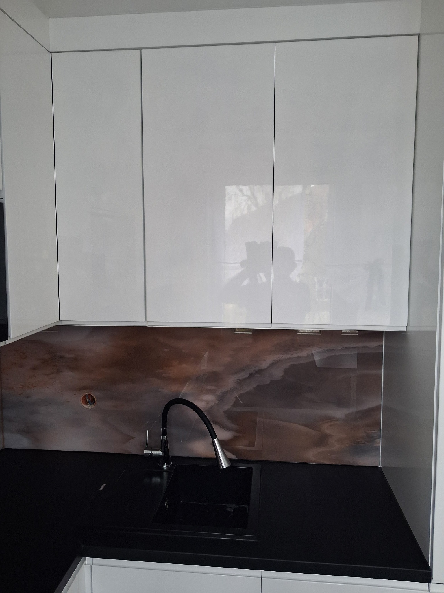 Białe, lakierowane szafki kuchenne wiszące nad czarnym blatem z wbudowanym zlewem i baterią. Na ścianie dekoracyjny panel imitujący marmur. Nowoczesny design.