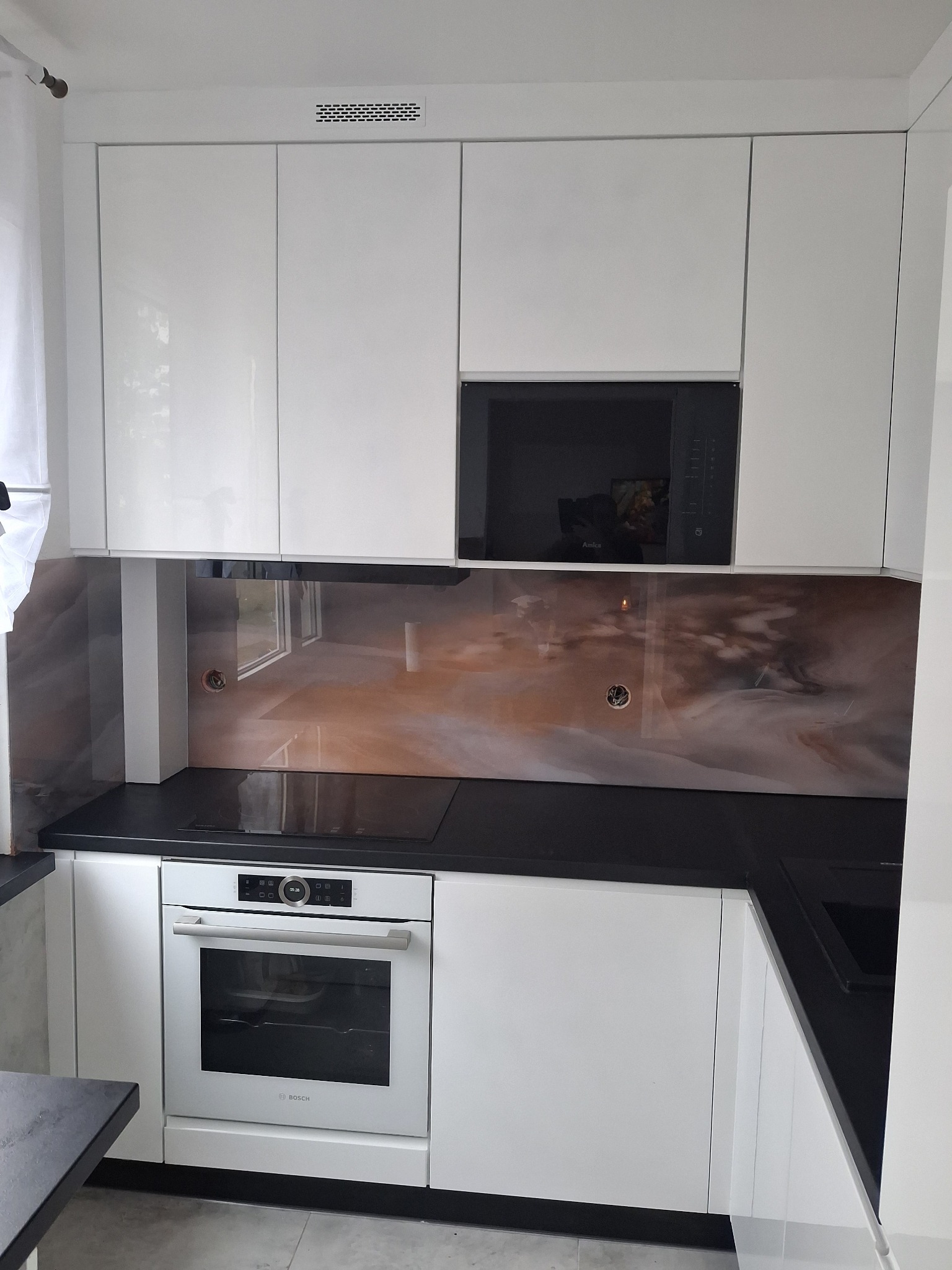 Biała, nowoczesna kuchnia z czarnym blatem i marmurowym panelem nad blatem. Widoczny piekarnik Bosch i mikrofalówka Amica. Górne szafki lakierowane na wysoki połysk.