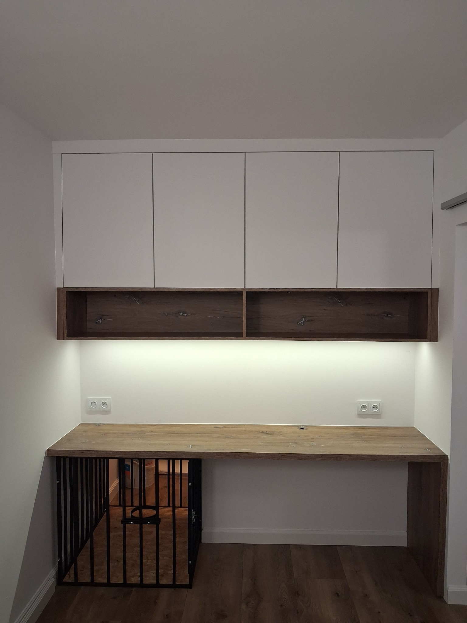 Białe szafki wiszące nad biurkiem z drewnianym blatem, podświetlane LED, z klatką dla psa wbudowaną pod blatem. Nowoczesny design, jasne wnętrze.