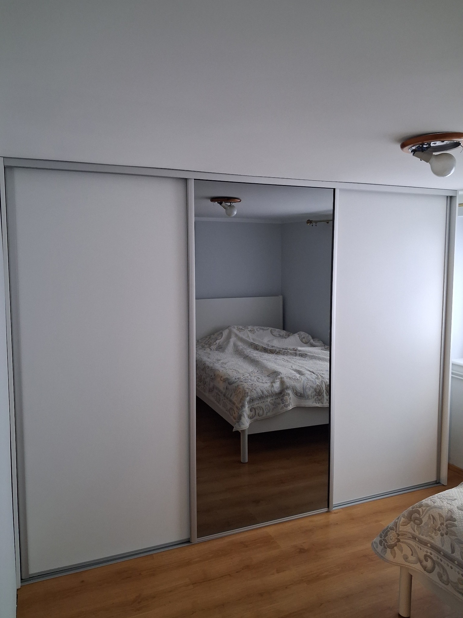 Biała szafa wnękowa z przesuwnymi drzwiami, częściowo lustrzanymi, odbijającymi wnętrze sypialni z łóżkiem. Minimalistyczny design, jasne wnętrze.