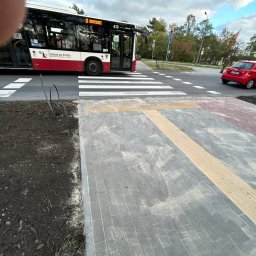 El-wer - Nowo wybudowany chodnik z płyt betonowych z żółtymi pasami dla osób niedowidzących, widoczny autobus miejski i samochody w tle.