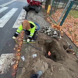 El-wer - Pracownik w odblaskowej kurtce mierzy żółtą miarką odległość przy wykopie obok chodnika, w tle minikoparka i stos kostki brukowej.