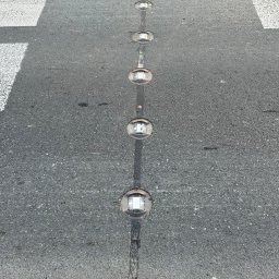 El-wer - Rząd wbudowanych w asfalt, okrągłych, odblaskowych świateł LED na przejściu dla pieszych, widocznych z góry, z elementami białego oznakowania poziomego.