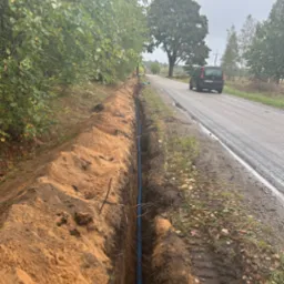 Wykop w ziemi z niebieskim kablem w peszlu, ułożonym wzdłuż drogi asfaltowej, w tle widoczny samochód i drzewa.