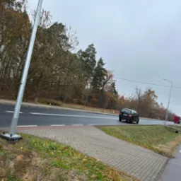 Słup oświetleniowy przy drodze, widoczne mocowanie do podłoża, w tle jesienny krajobraz i przejeżdżające samochody.