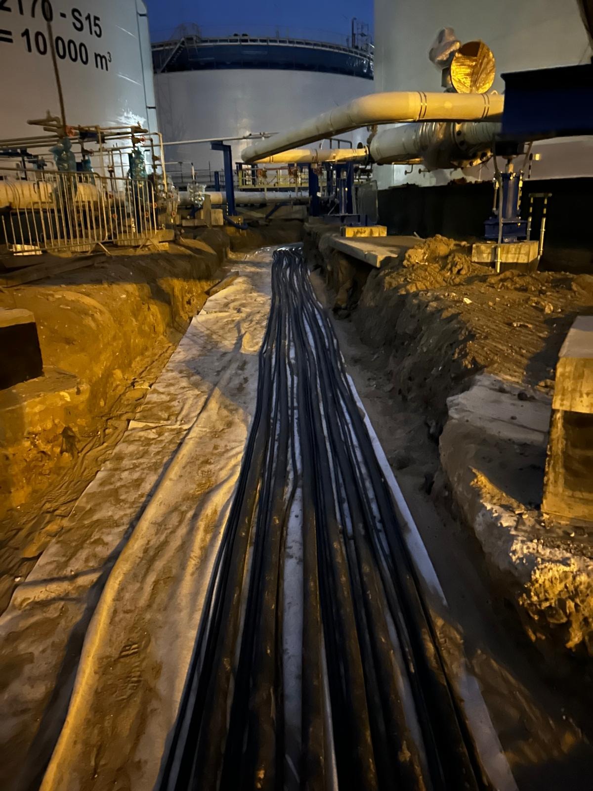 Wykop z ułożonymi równolegle kablami w osłonie, widoczne fragmenty instalacji przemysłowej i zbiorniki w tle, nocna sceneria.