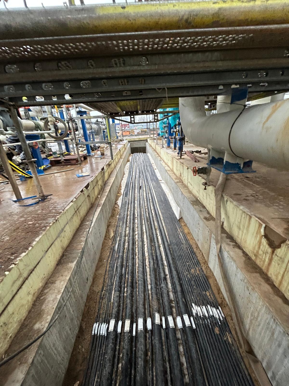 Wykop z ułożonymi kablami elektrycznymi w korycie betonowym, widoczne etykiety identyfikacyjne na kablach, w tle rurociągi i konstrukcja stalowa.