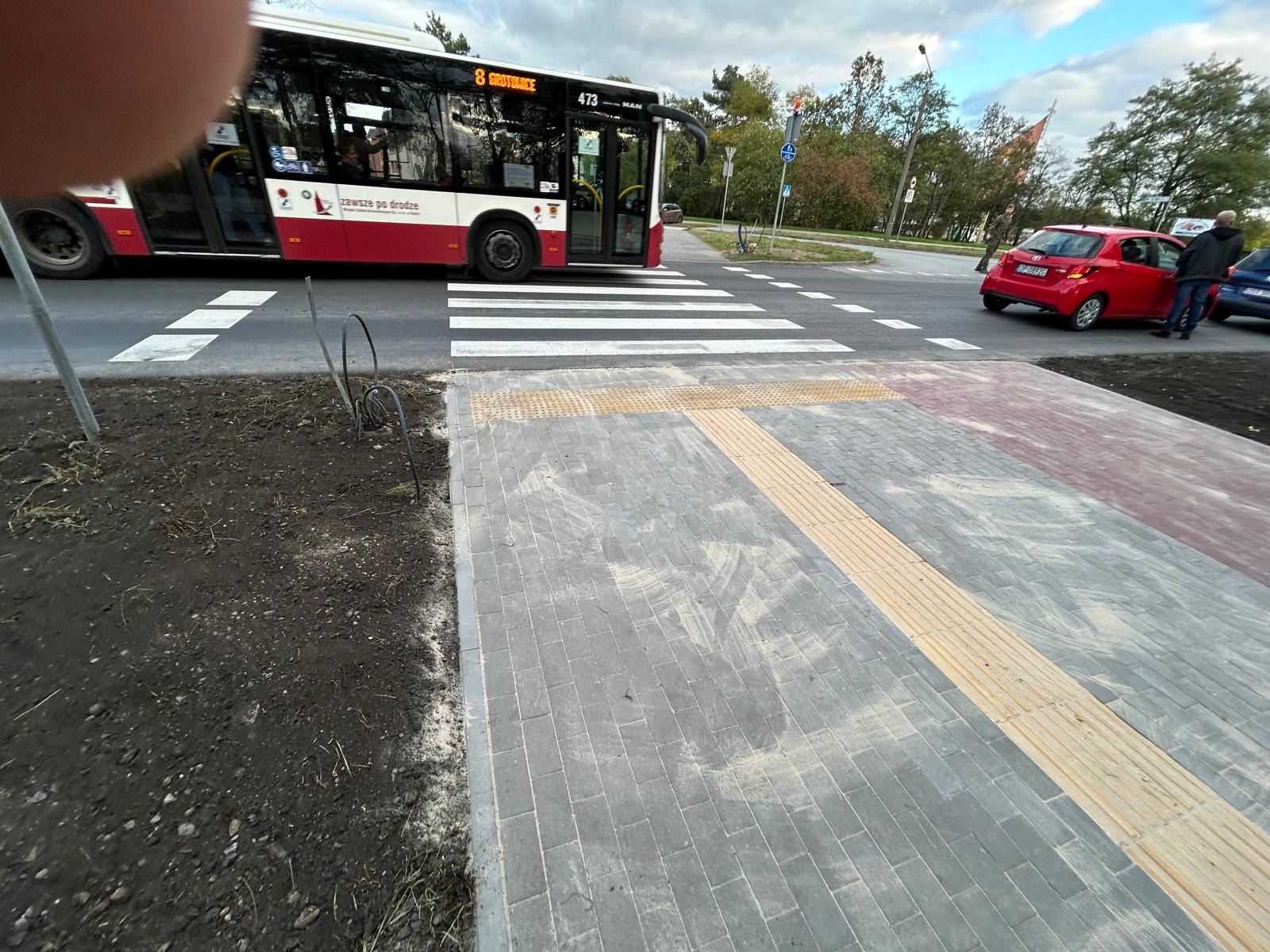 Nowo wybudowany chodnik z płyt betonowych z żółtymi pasami dla osób niedowidzących, widoczny autobus miejski i samochody w tle.