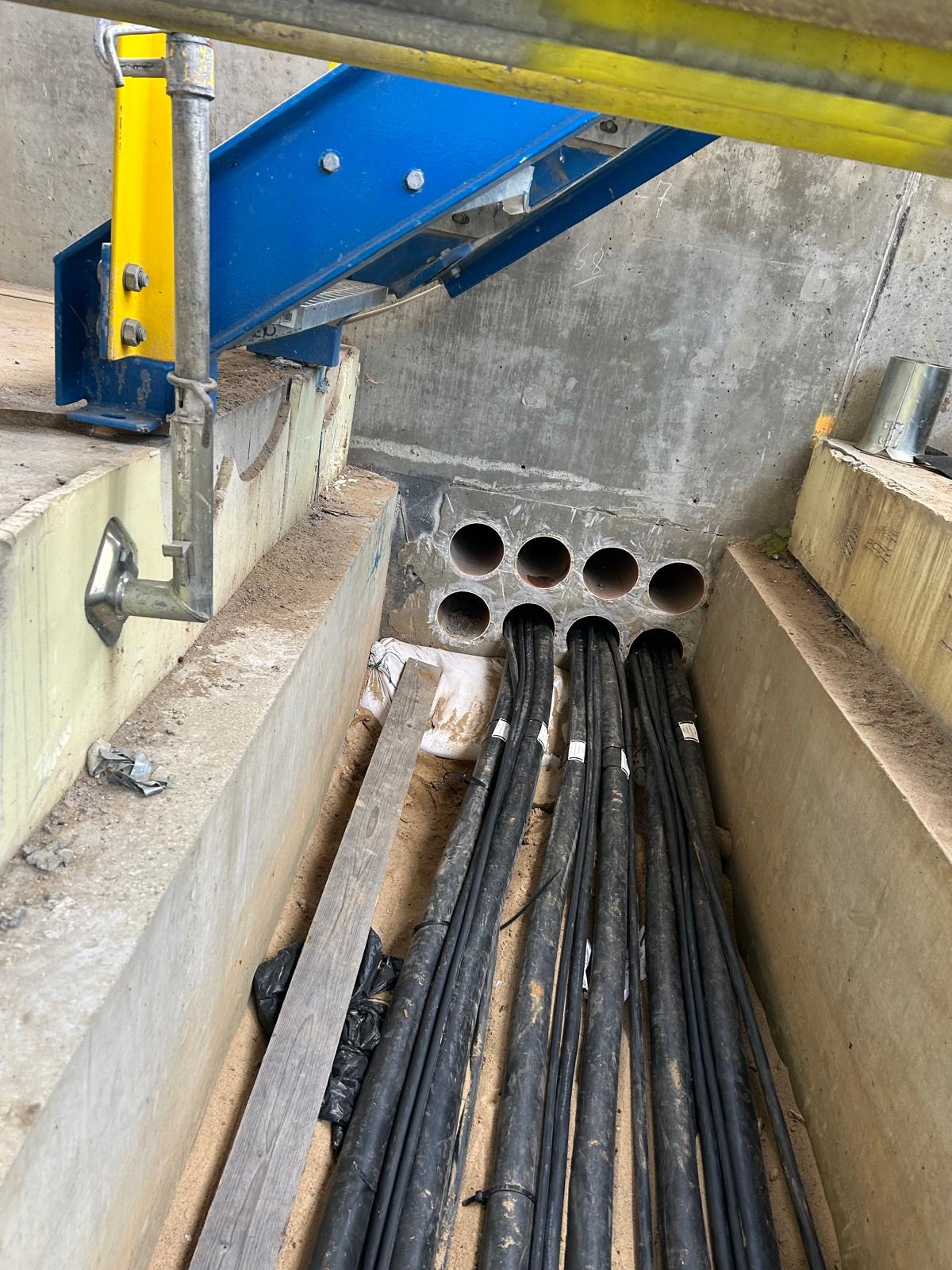 Wykop z betonowymi ścianami, przez otwory w ścianie przechodzą grube czarne kable elektryczne, widoczna konstrukcja stalowa nad wykopem.